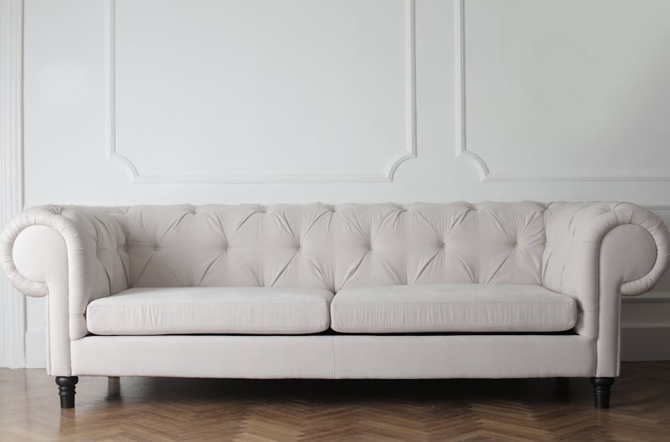 A white, tufted sofa with rolled arms and dark wooden legs positioned against a white wall with decorative paneling. The sofa's upholstery appears clean and well-maintained, set on a light hardwood fl