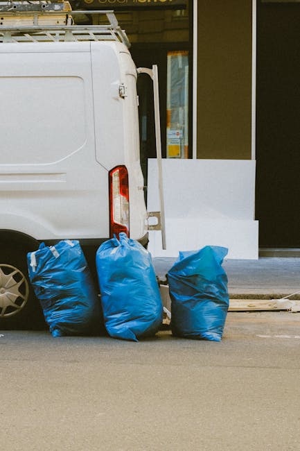 A large green wheeled waste bin marked with the number 53 and the words 'JUN BESAR' positioned on a sidewalk next to a storage rack holding cleaning supplies and tools. The scene is illuminated by nat